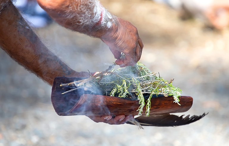photo of a First Nations man's hands holding a feather, some bark and some leaves which he is trying to burn